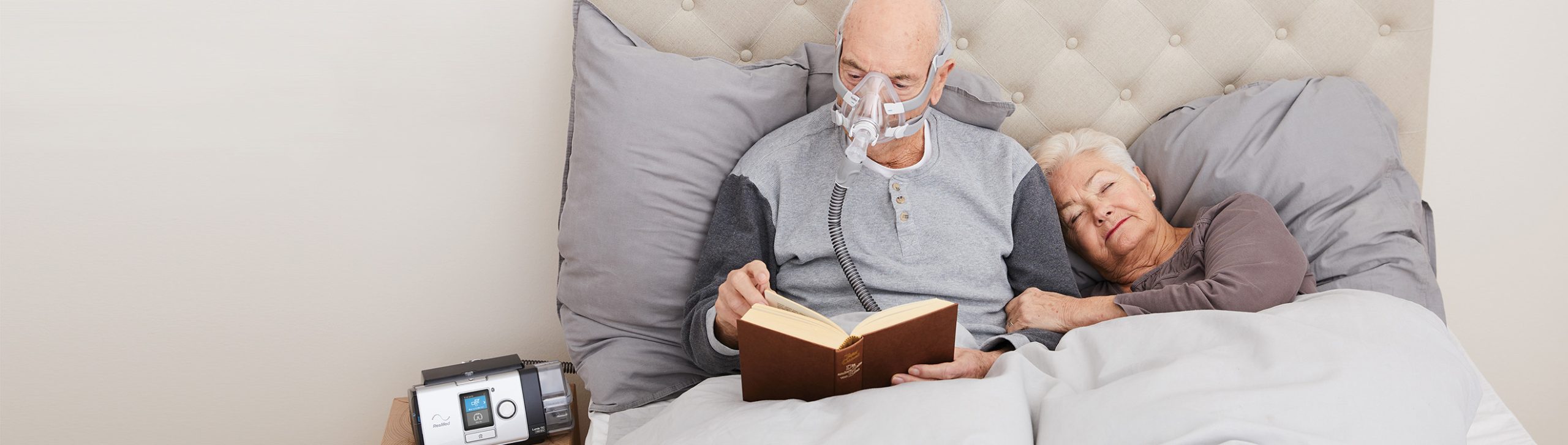 Ein älterer Mann mit Atemmaske liest im Bett ein Buch, während eine ältere Frau neben ihm liegt und sich ausruht.