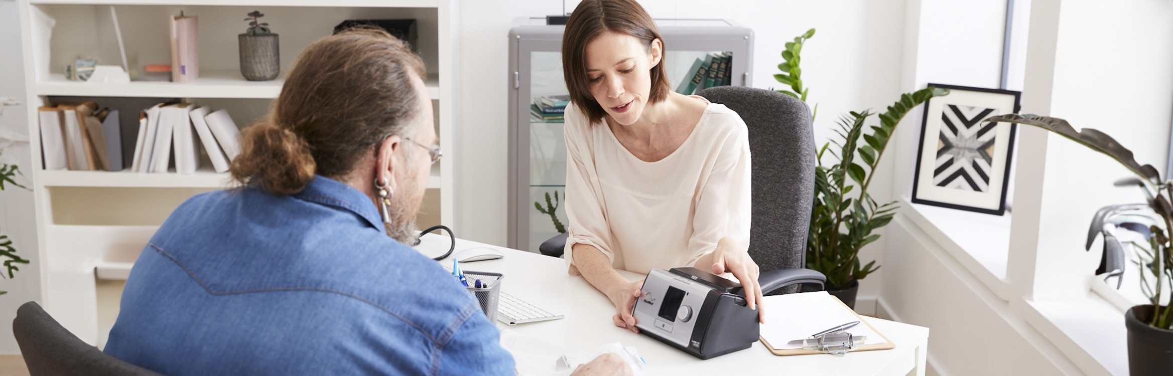 Eine Ärztin erklärt einem Patienten die Funktionsweise eines CPAP-Geräts während eines Beratungsgesprächs.