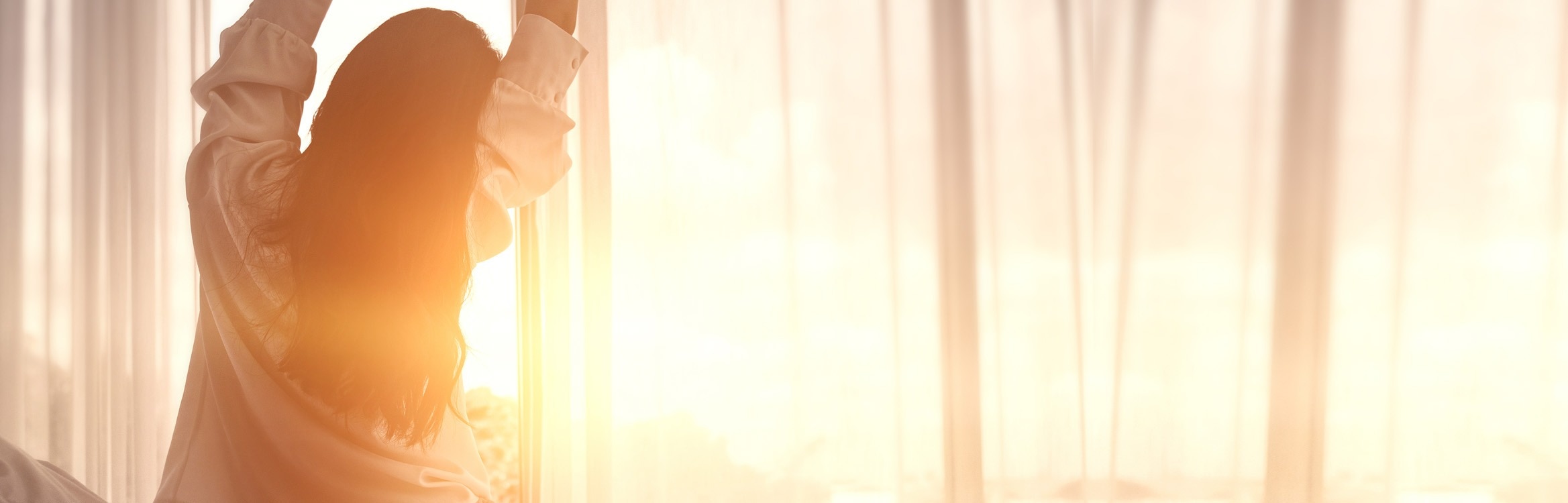 Frau streckt sich morgens vor einem Fenster mit Sonnenaufgang.