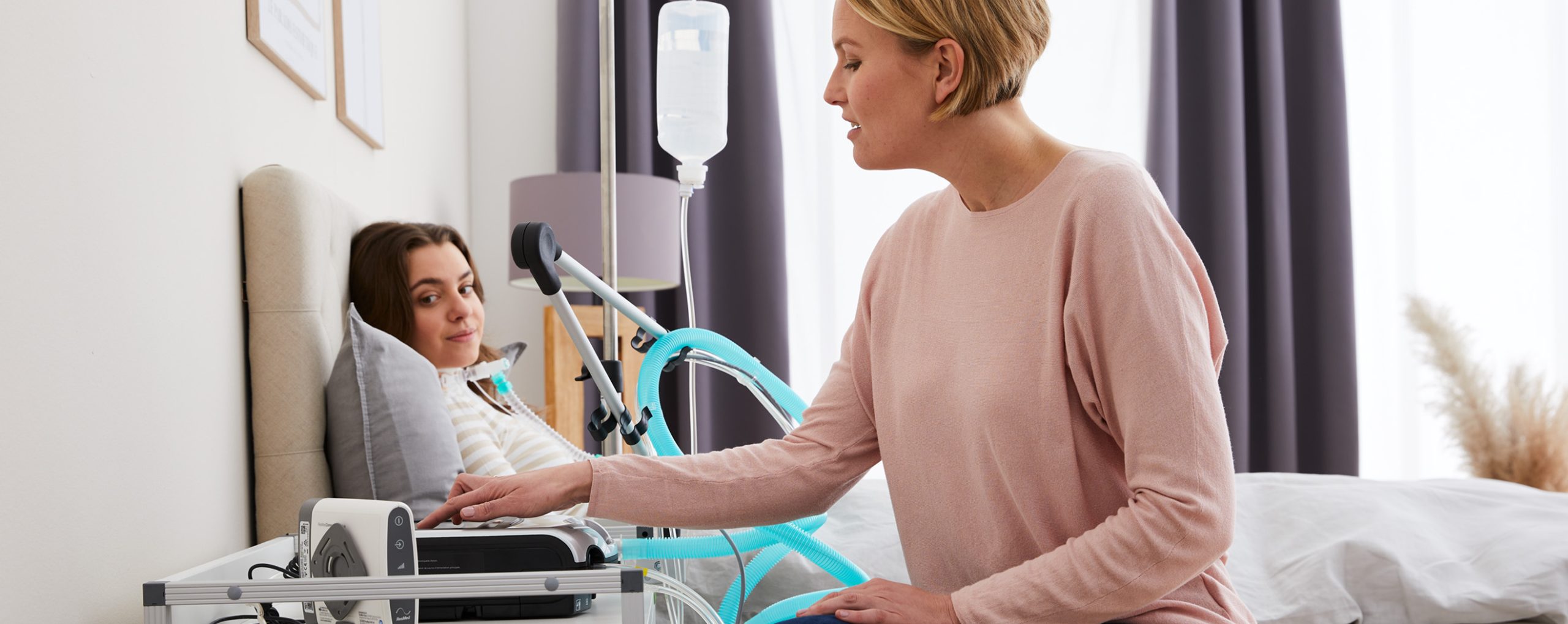 Eine Fachkraft bedient ein Beatmungsgerät, während eine junge Patientin im Bett liegt und eine nicht-invasive Beatmungstherapie erhält.