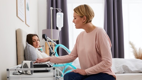 Eine Fachkraft bedient ein Beatmungsgerät, während eine junge Patientin im Bett liegt und eine nicht-invasive Beatmungstherapie erhält.