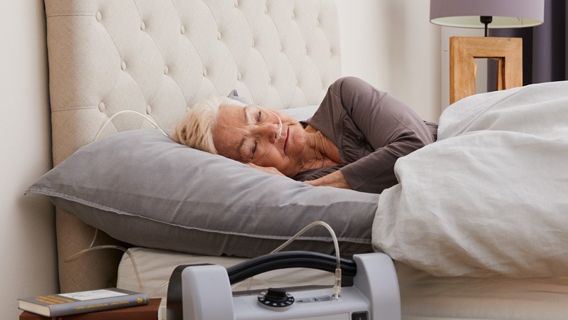 Eine ältere Frau mit Sauerstoffschlauch schläft friedlich in einem Bett, während ein Sauerstoffgerät neben dem Bett steht.
