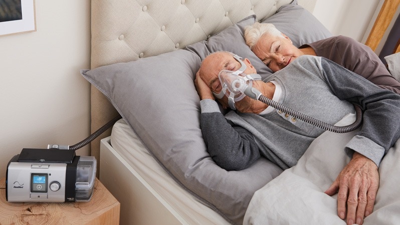 Älteres Paar liegt im Bett, der Mann trägt eine Atemmaske, verbunden mit einem Beatmungsgerät auf dem Nachttisch.